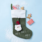 An green Christmas stocking with a white cuff, featuring a 3D Santa face applique, white 'HO! HO! HO!' text, and a green hanging loop. The stocking is styled on a light blue background with scattered white faux snow, candy canes, gingerbread cookies, and a small red-and-white striped ornament.