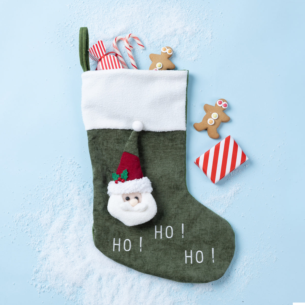 An green Christmas stocking with a white cuff, featuring a 3D Santa face applique, white 'HO! HO! HO!' text, and a green hanging loop. The stocking is styled on a light blue background with scattered white faux snow, candy canes, gingerbread cookies, and a small red-and-white striped ornament.