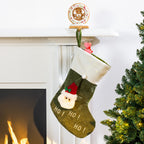 A green Christmas stocking with a white cuff, featuring a plush Santa face and the text 'HO! HO! HO!' hanging by a green loop in front of a white fireplace mantel. A small gingerbread ornament sits on the mantel above, and a Christmas tree is visible on the right.