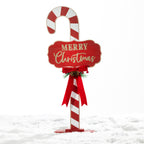 A festive Christmas yard sign shaped like a candy cane, featuring a red and white striped cane body, a red plaque with gold glittery 'MERRY Christmas' text, and a red ribbon bow with small green pine accents, set in white faux snow against a white background.