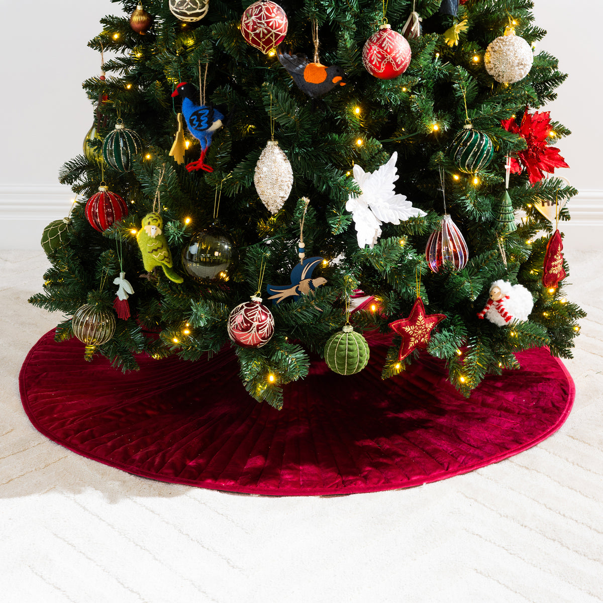 Christmas Wishes Red Channel Velvet Tree Skirt displayed beneath a Christmas tree adorned with colourful ornaments and warm festive lights.