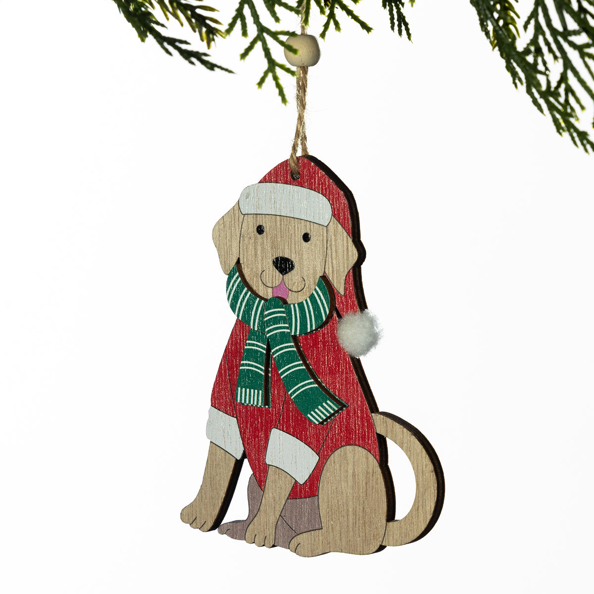 A wooden Christmas ornament shaped like a sitting dog wearing a red sweater, green striped scarf, and red Santa hat, hanging from a jute string under pine branches against a white background.