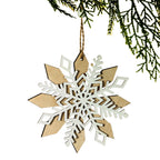 A wooden snowflake Christmas ornament with layered natural wood and white laser-cut details, hanging from a twine string under a green pine branch against a white background.