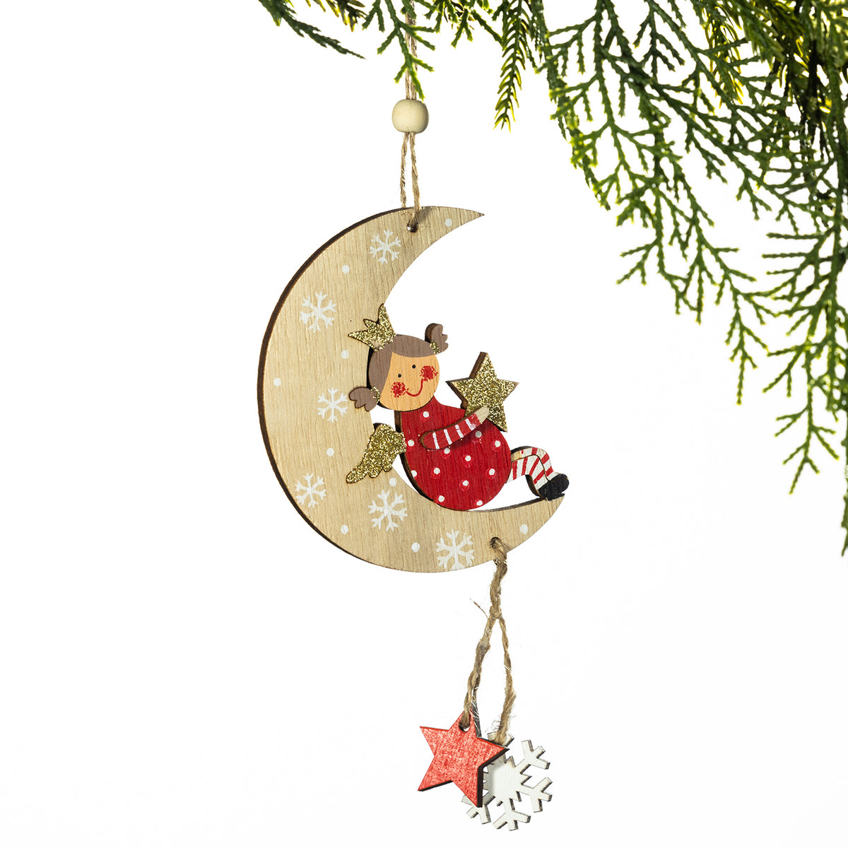 A wooden Christmas ornament shaped like a crescent moon with a sleeping angel figure in red and white, decorated with white snowflakes. A small hanging star and white snowflake charm dangle below, suspended by twine from a pine branch against a white background.