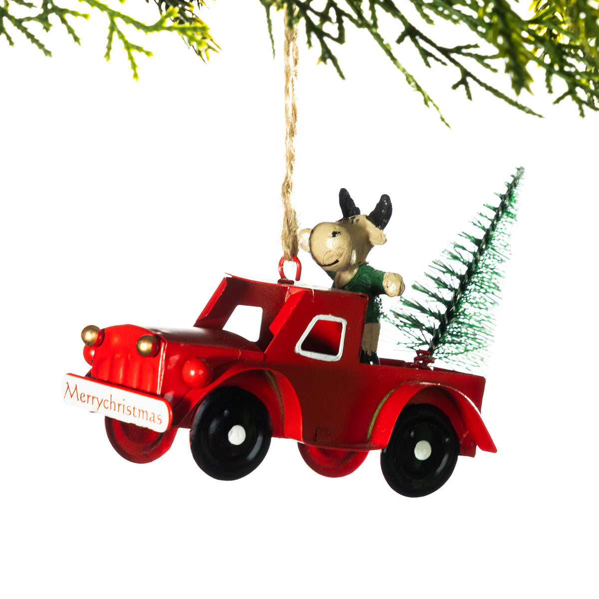 A Christmas ornament shaped like a red pickup truck carrying a small green tree and a cow figurine, hanging from a string against a white background with evergreen branches at the top.