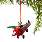 A Christmas ornament shaped like a small red airplane with green wings, a white propeller, and a small Santa-clad figure in the cockpit, hanging from an orange ribbon beneath evergreen branches on a white background.