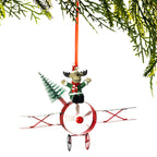 A Christmas ornament shaped like a reindeer riding a red and white biplane, with a small green tree and an orange hanging ribbon, shown against a white background with pine branches at the top.
