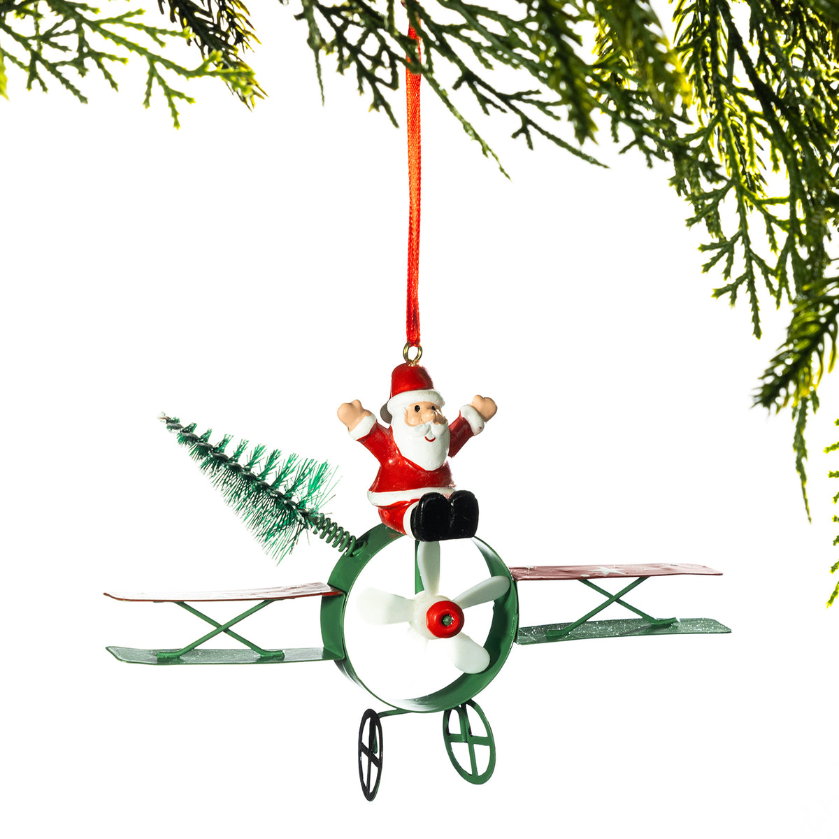 A Christmas ornament shaped like a small green airplane with Santa Claus sitting on top, hanging from a red ribbon against a white background with pine branches framing the top.