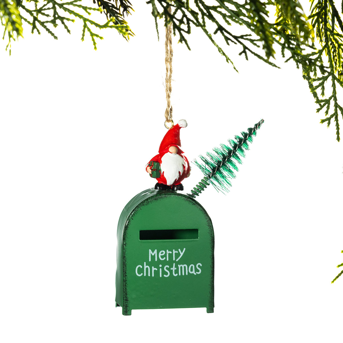 A green mailbox Christmas ornament hanging from a tree branch, featuring a small gnome on top, a miniature green tree, and 'Merry Christmas' written on the front.