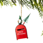 A Christmas ornament shaped like a red mailbox with a gnome figure and a small tree on top, hanging from a gold string beneath evergreen branches against a white background.