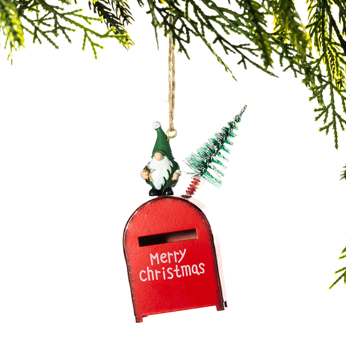 A Christmas ornament shaped like a red mailbox with a gnome figure and a small tree on top, hanging from a gold string beneath evergreen branches against a white background.