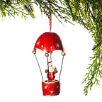 A Christmas hot air balloon ornament featuring a small Santa Claus figure in a red basket beneath a dome-shaped red canopy with gold accents, hanging against a white background with pine branches framing the top and right edges.