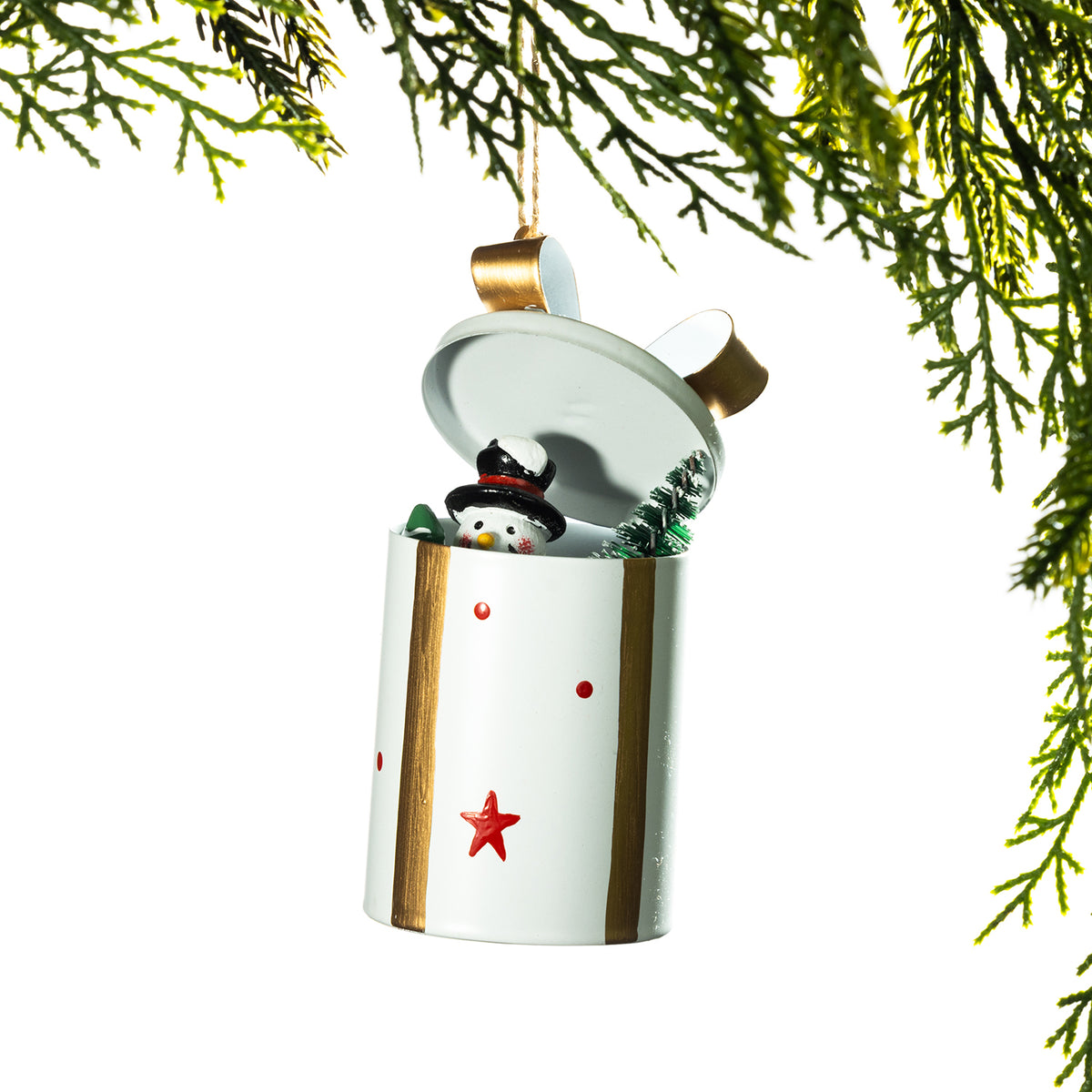 A white cylindrical Christmas ornament shaped like a gift box with a hinged lid, decorated with red stars and dots and gold side accents. A small snowman figure peeks out from inside, and green pine branches frame the top and sides against a white background.