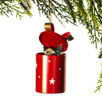 A red cylindrical Christmas ornament shaped like an open gift box, with white star and dot cutouts, a gold ribbon loop, and a small holiday figurine peeking out. The ornament is shown hanging from a green pine branch against a white background.