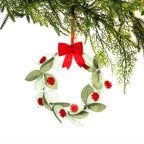 A green felt Christmas wreath featuring layered sage and olive leaves with red pōhutukawa flower details and a bright red bow, hanging from a Christmas tree branch against a white background.