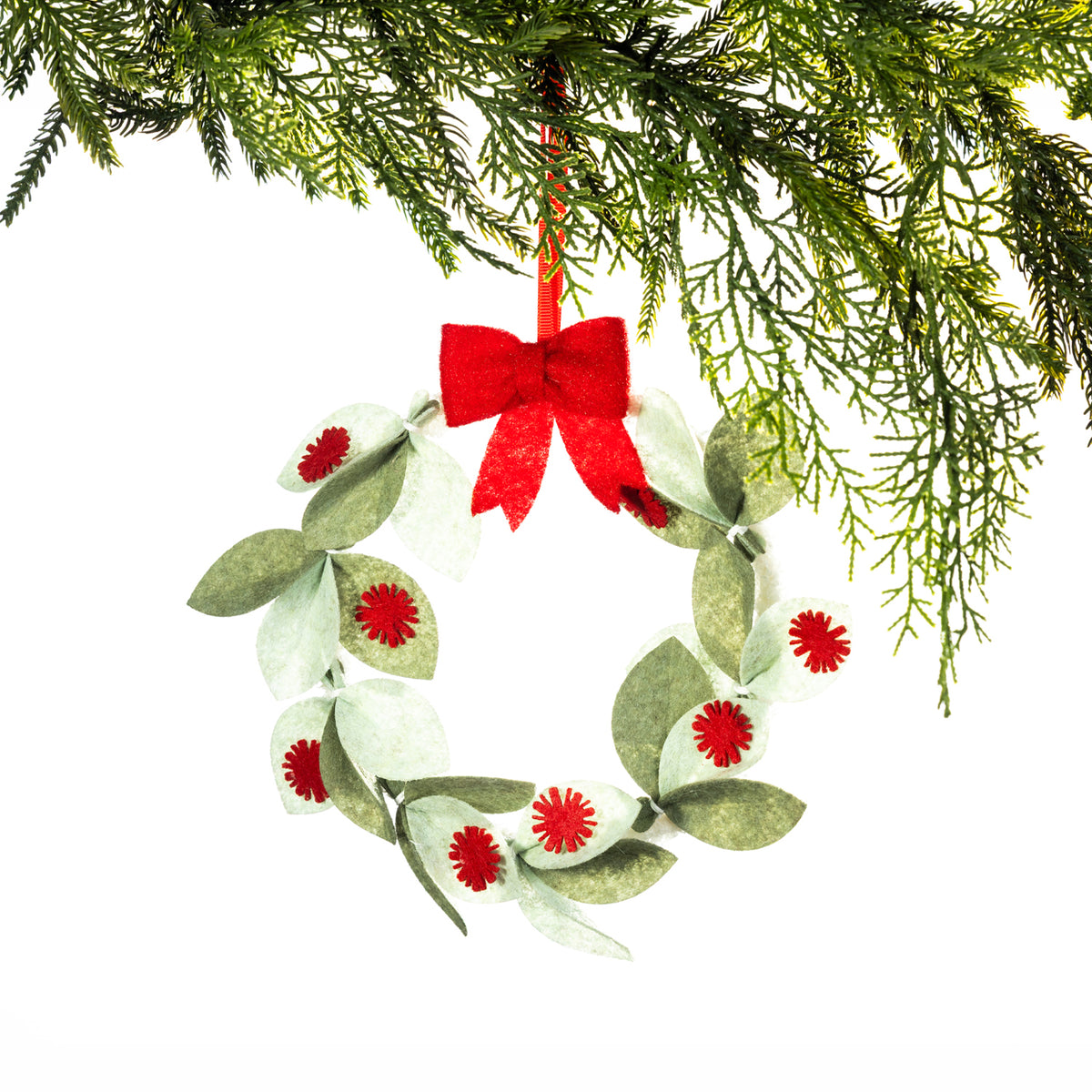 A green felt Christmas wreath featuring layered sage and olive leaves with red pōhutukawa flower details and a bright red bow, hanging from a Christmas tree branch against a white background.