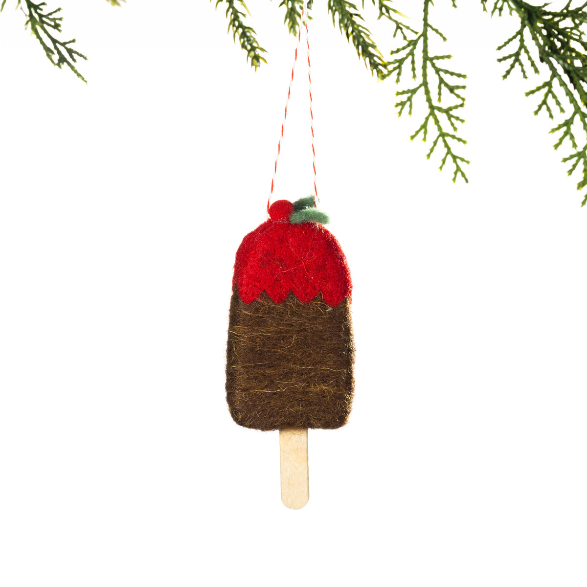 A brown felt chocolate popsicle decoration featuring a red topping, green holly leaves and a wooden stick, hanging from a Christmas tree branch against a white background.