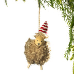 A fluffy sheep-shaped hanging ornament with curly tan wool, a wooden face, white ears, and a red-and-white striped cone hat, suspended by a string against a white background with green pine branches at the top right.