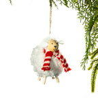 A fluffy white sheep Christmas decoration featuring a smiling wooden face, soft curly wool, wooden legs and a red and white striped scarf, hanging from a Christmas tree branch.