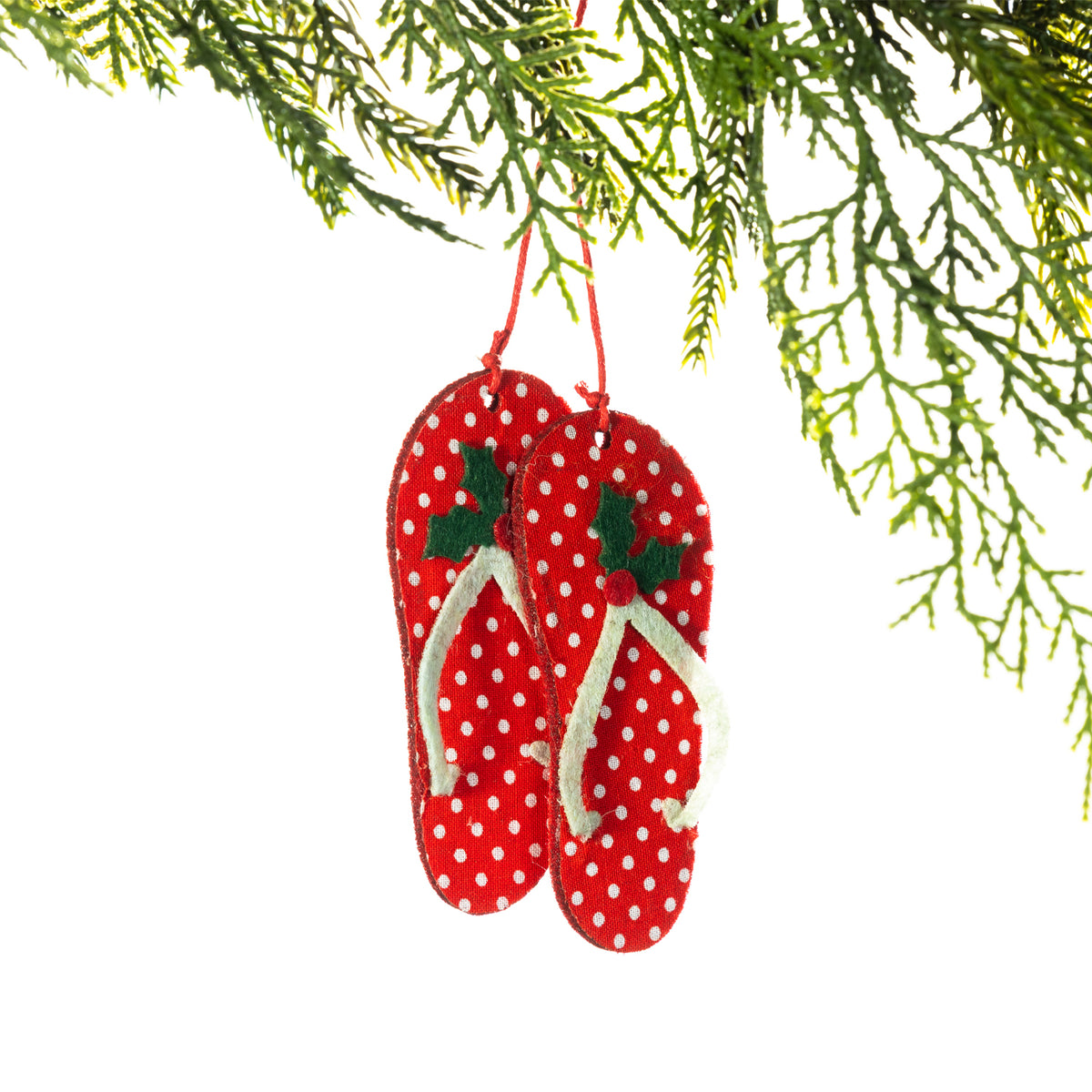 A pair of red felt jandals featuring white polka dots, white straps and green holly leaf detailing, hanging from a Christmas tree branch against a white background.