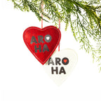 Two heart-shaped felt Christmas decorations in red and white, featuring bold black Aroha lettering, contrasting heart details and hanging loops, displayed on a Christmas tree branch.