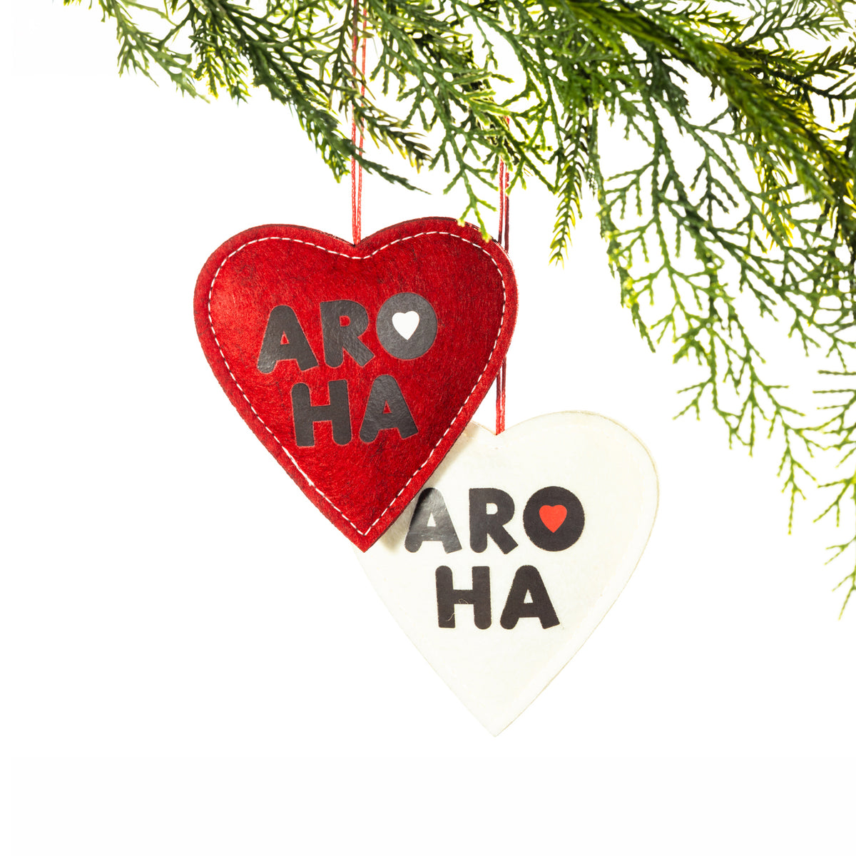 Two heart-shaped felt Christmas decorations in red and white, featuring bold black Aroha lettering, contrasting heart details and hanging loops, displayed on a Christmas tree branch.