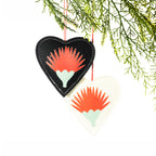 Two heart-shaped felt Christmas decorations in black and cream, featuring red pōhutukawa flower designs, contrasting stitching and red hanging loops, displayed on a Christmas tree branch.