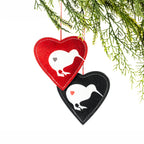 Two heart-shaped felt Christmas decorations in red and black, featuring white kiwi bird silhouettes, contrasting heart details and stitched edging, hanging from a Christmas tree branch.