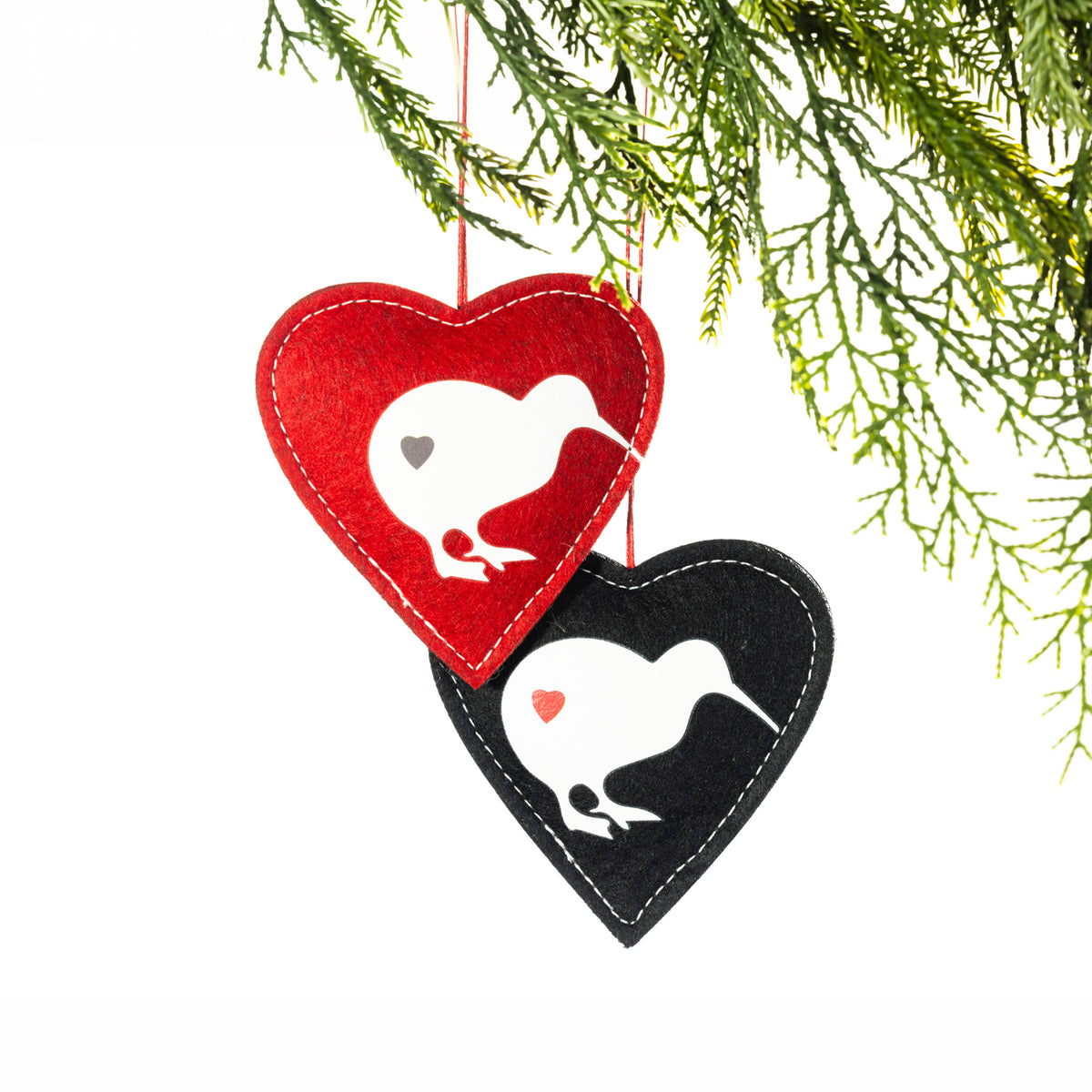 Two heart-shaped felt Christmas decorations in red and black, featuring white kiwi bird silhouettes, contrasting heart details and stitched edging, hanging from a Christmas tree branch.