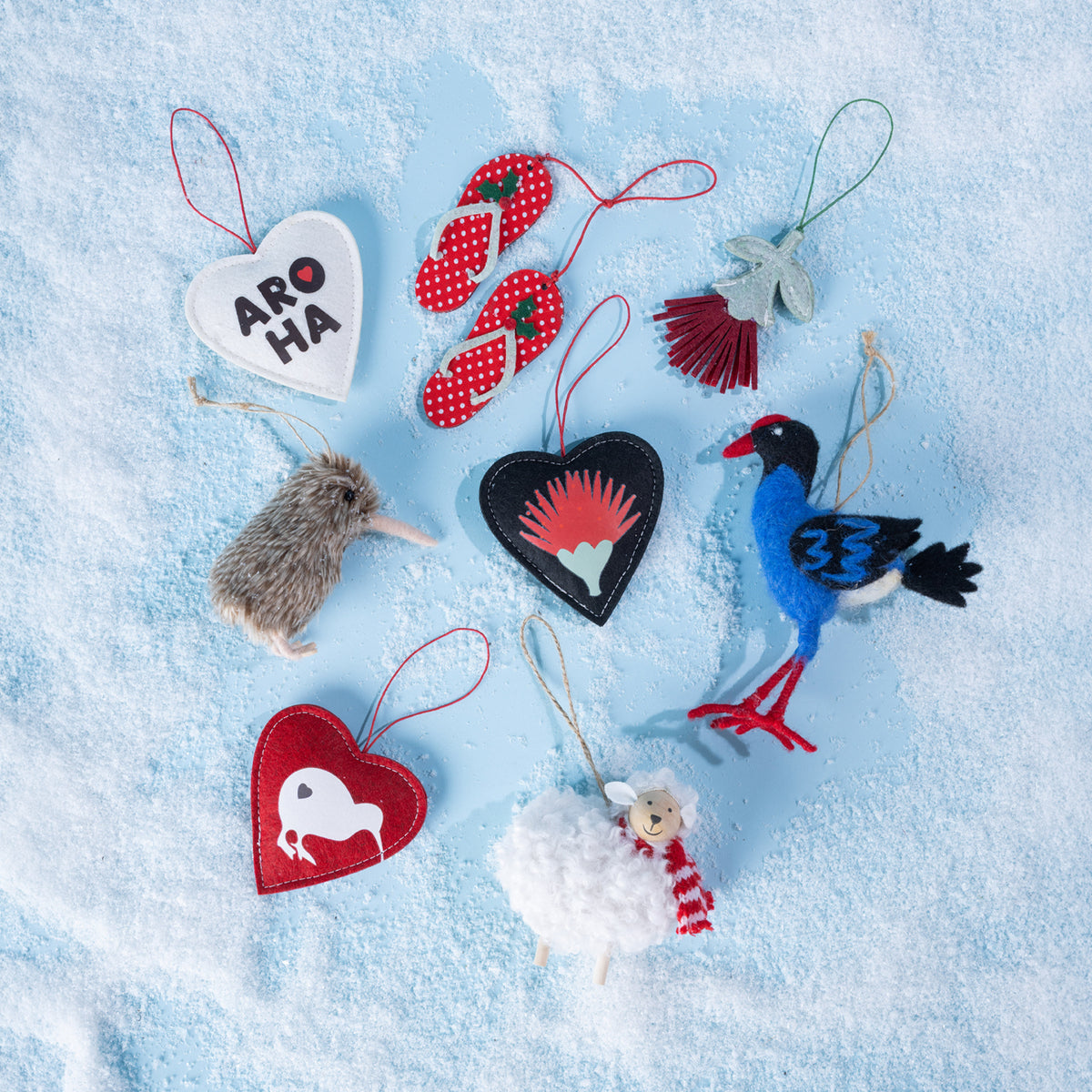 A collection of colourful New Zealand-inspired felt Christmas decorations featuring kiwi birds, pōhutukawa flowers, red jandals, a sheep and decorative hearts, displayed on a snowy light blue background.