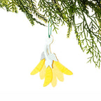 A yellow felt kōwhai flower Christmas decoration featuring layered yellow petals, a pale green stem and leaves, hanging from a Christmas tree branch against a white background.