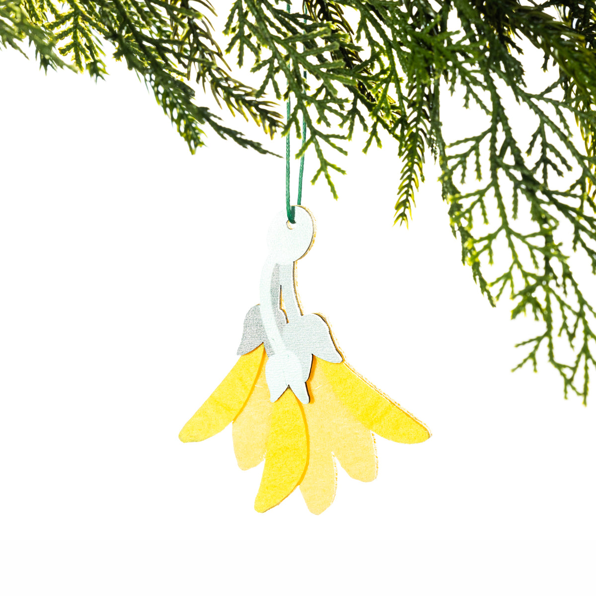 A yellow felt kōwhai flower Christmas decoration featuring layered yellow petals, a pale green stem and leaves, hanging from a Christmas tree branch against a white background.