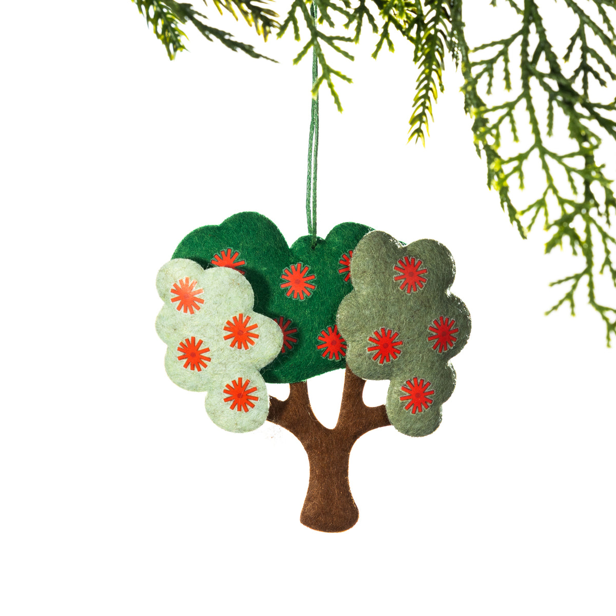 A felt pōhutukawa tree Christmas decoration featuring layered green foliage in different shades, bright red flower details and a brown tree trunk, hanging from a Christmas tree branch against a white background.