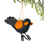 A felt bird ornament in dark charcoal gray with a bright orange chest and beak, hanging from a thin string beneath evergreen branches against a white background.