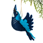 A felt bird ornament in dark navy blue and bright turquoise blue, hanging from a thin string against a white background with green pine branches at the top right.