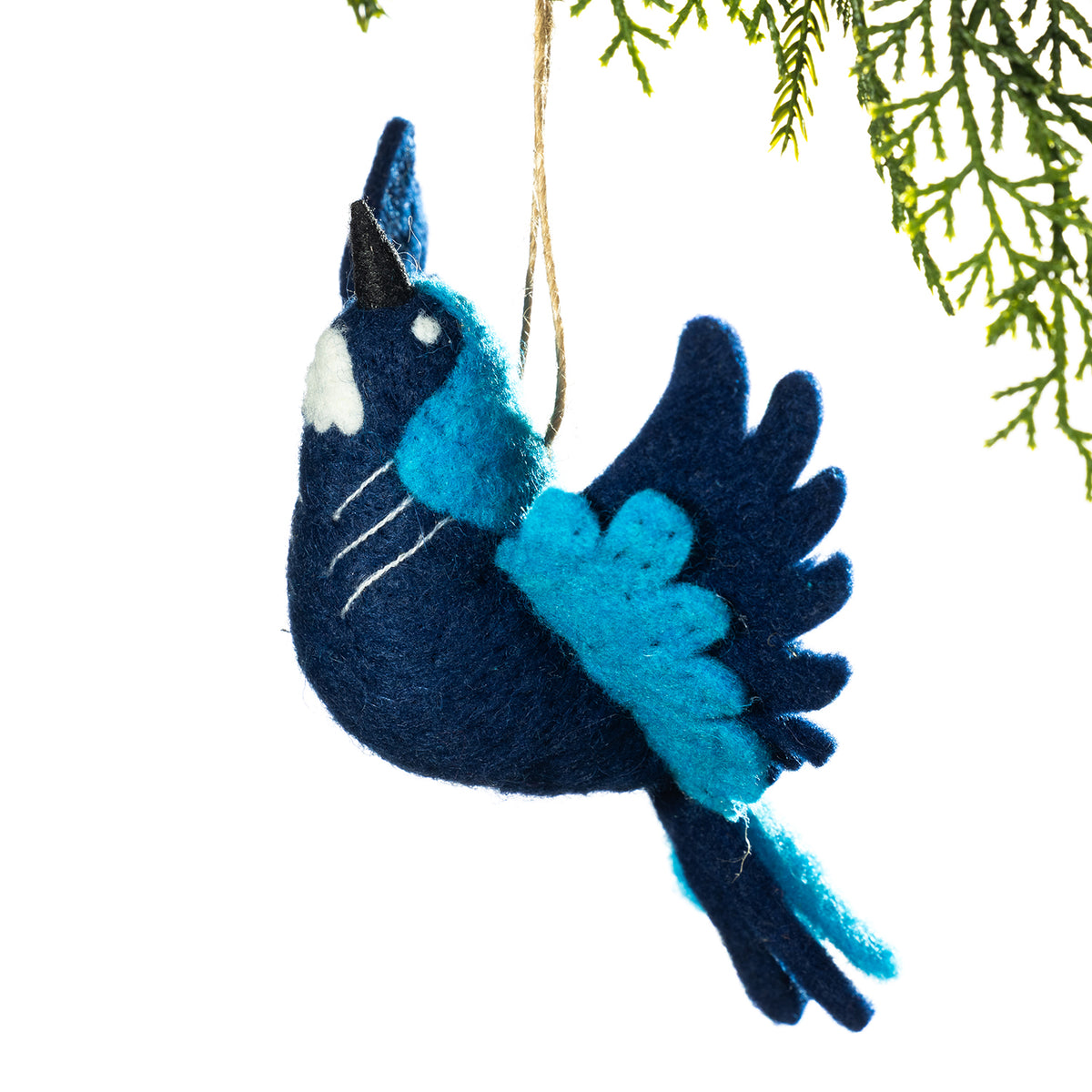 A felt bird ornament in dark navy blue and bright turquoise blue, hanging from a thin string against a white background with green pine branches at the top right.