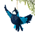 A felt bird ornament in dark navy blue and bright turquoise blue, hanging from a thin string against a white background with green pine branches at the top right.