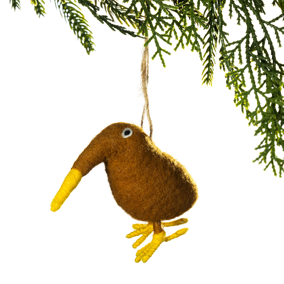 A brown felt bird ornament with a yellow beak and feet, hanging from a string beneath green pine branches against a white background.
