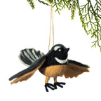 A felt bird ornament, shaped like a small black, white, and tan bird with spread wings, hanging from a string against a white background with green pine branches at the top.