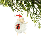 A fluffy white sheep Christmas decoration featuring a smiling wooden face, red Santa hat, wooden legs and a red present with black ribbon detailing, hanging from a Christmas tree branch against a white background.
