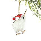 A fluffy white kiwi Christmas decoration featuring soft feather-like fur, a long wooden beak, wooden feet and a red Santa hat with a small bell, hanging from a Christmas tree branch against a white background.