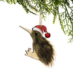 A fluffy brown kiwi Christmas decoration featuring soft natural brown fur, a long wooden beak, wooden feet and a red Santa hat with a small bell, hanging from a Christmas tree branch against a white background.