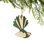 A wooden pīwakawaka Christmas decoration featuring a green and natural wood colourway, layered wings and a distinctive fan-shaped tail, hanging from a Christmas tree branch against a white background.
