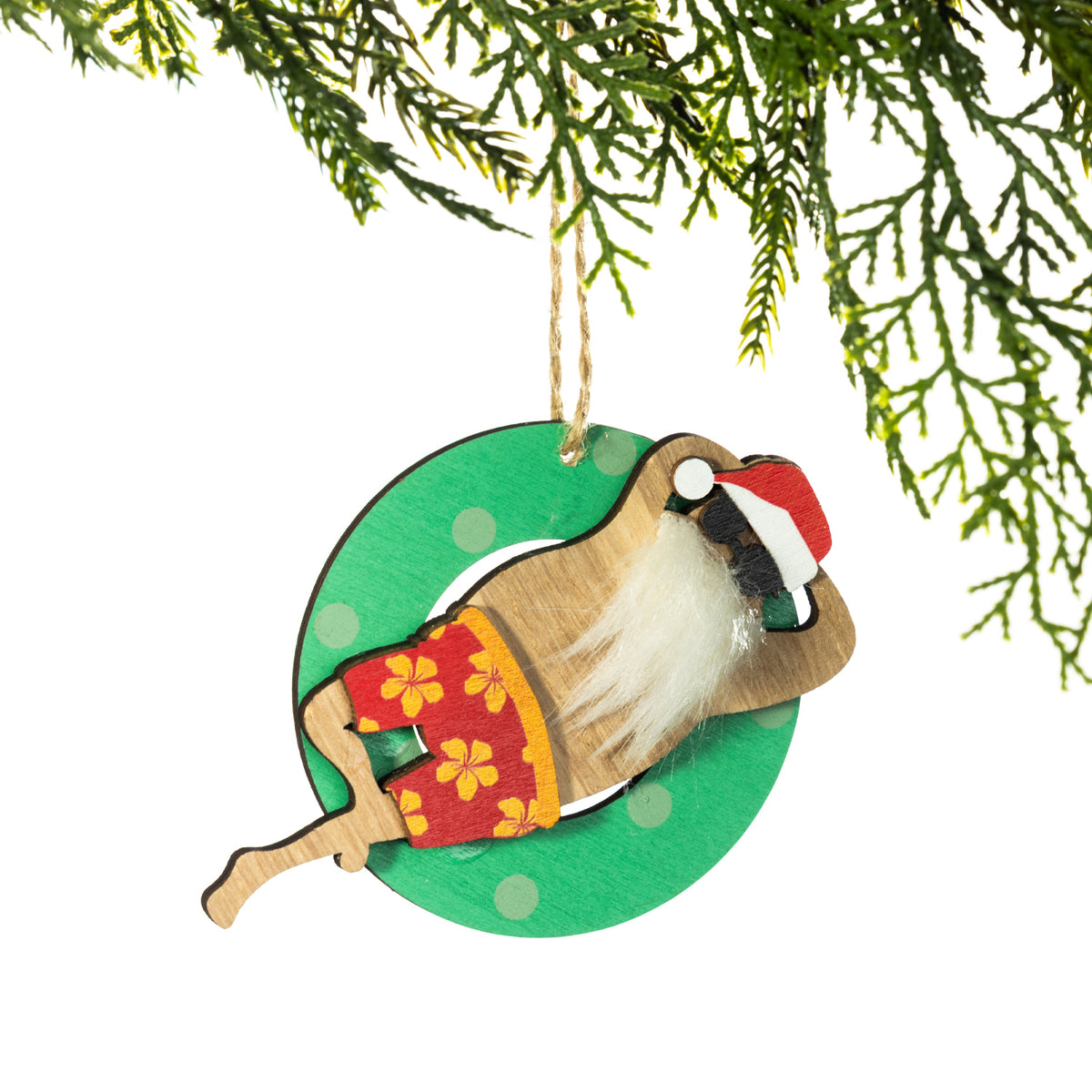 A wooden Santa Christmas decoration featuring a fluffy white beard, black sunglasses, red floral board shorts and a Santa hat, relaxing on a green polka dot pool float, hanging from a Christmas tree branch against a white background.