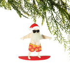A wooden surfing Santa Christmas decoration featuring a fluffy white beard, black sunglasses, red floral board shorts and a Santa hat, standing on a red surfboard, hanging from a Christmas tree branch against a white background.
