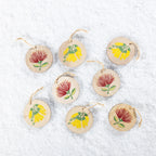 A set of eight round wooden Christmas decorations featuring colourful red pōhutukawa and yellow kōwhai flower designs, finished with natural jute hanging loops and arranged on a snowy white background.