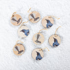 A set of eight round wooden Christmas decorations featuring native New Zealand tūī and pīwakawaka bird designs in blue, black and grey, finished with natural jute hanging loops and displayed on a snowy white background.