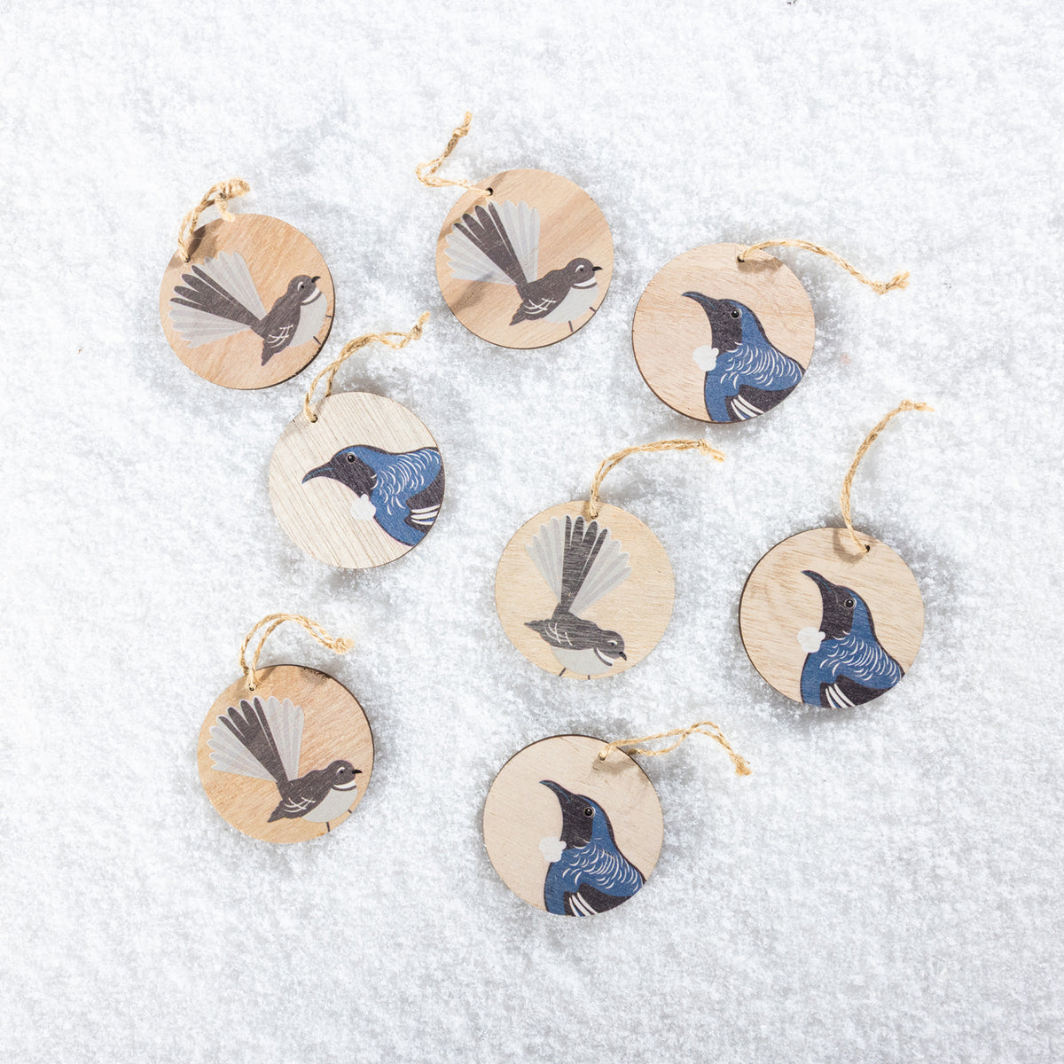 A set of eight round wooden Christmas decorations featuring native New Zealand tūī and pīwakawaka bird designs in blue, black and grey, finished with natural jute hanging loops and displayed on a snowy white background.