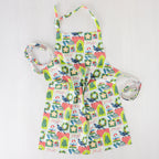 Christmas Wishes Kiwi Christmas Apron laid flat, featuring a colourful New Zealand-inspired festive print with Christmas trees, wreaths, ornaments, gifts and native birds.