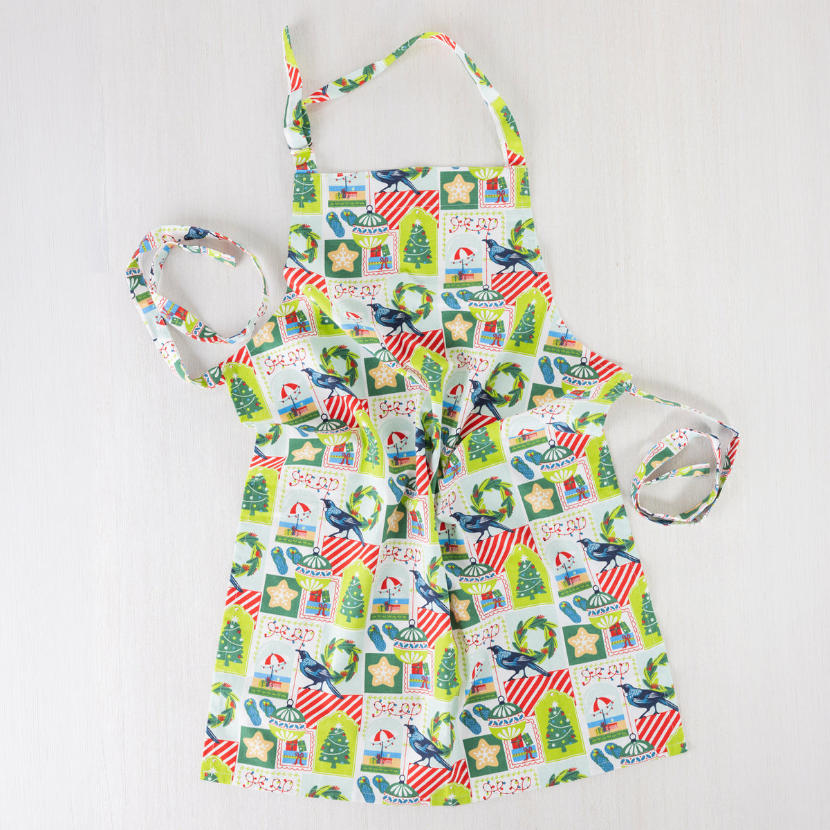 Christmas Wishes Kiwi Christmas Apron laid flat, featuring a colourful New Zealand-inspired festive print with Christmas trees, wreaths, ornaments, gifts and native birds.